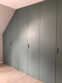 Armoire sous pente: chaque toit a une inclinaison différente, c&rsquo;est pourquoi nous proposons notre configurateur, qui s&rsquo;adapte parfaitement à vos mesures pour créer un design d&rsquo;armoire pratique et fonctionnel, personnalisé selon vos besoins. Tout est soigneusement dissimulé derrière des portes aux couleurs personnalisées. Cette armoire est installée dans la maison de notre client à Vienne, Autriche – FB 85 Oval Room Blue laqué mat avec poignées intégrées S1.